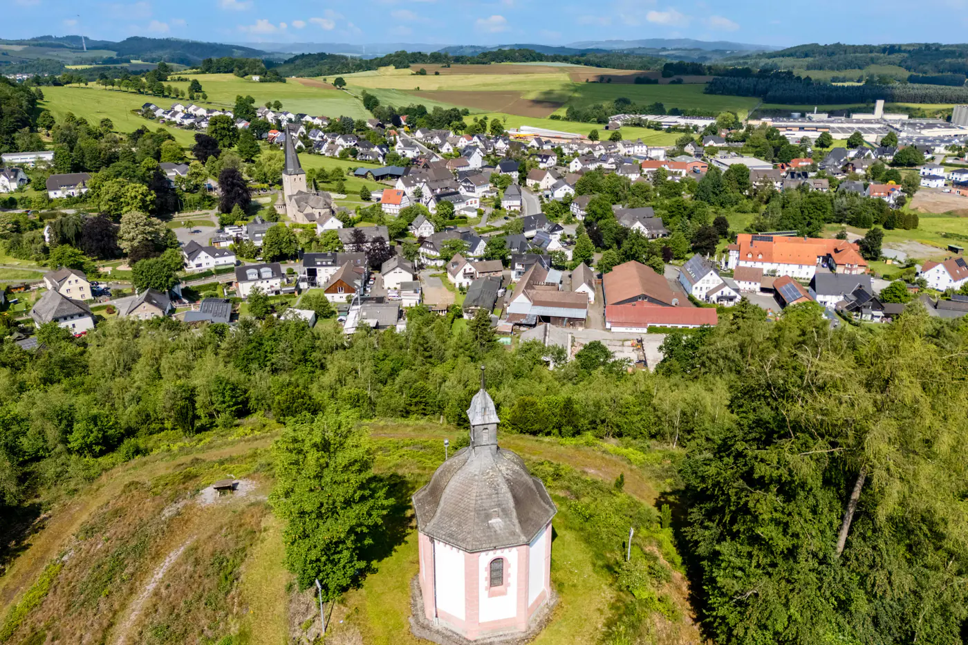 Die Rehbergkapelle auf dem bewaldeten Höhenrücken über Stockum, dahinter das Dorf und die Felder des Sauerlands